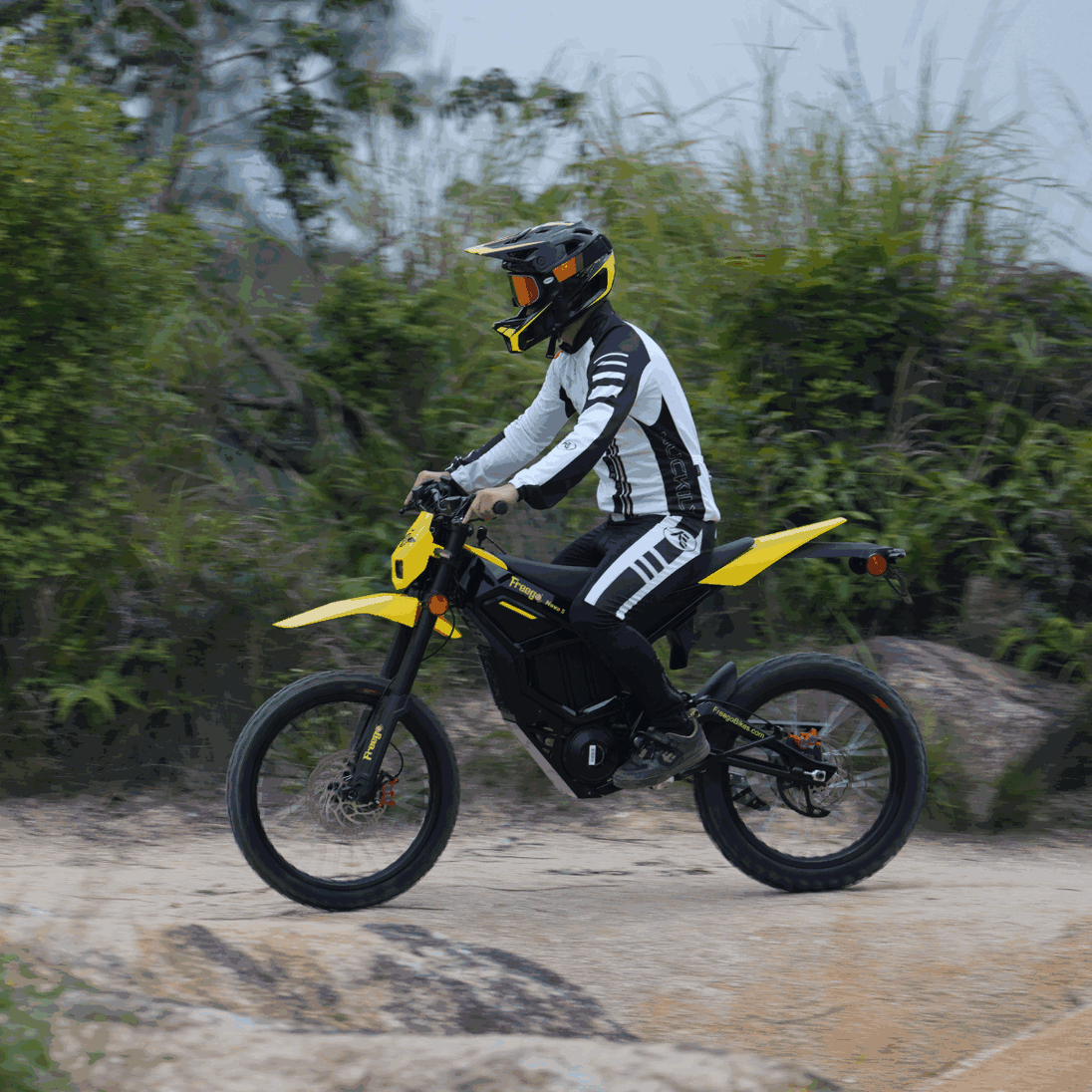 Rider on Freego Nova 5 electric dirt bike riding past dense greenery on an off-road trail.