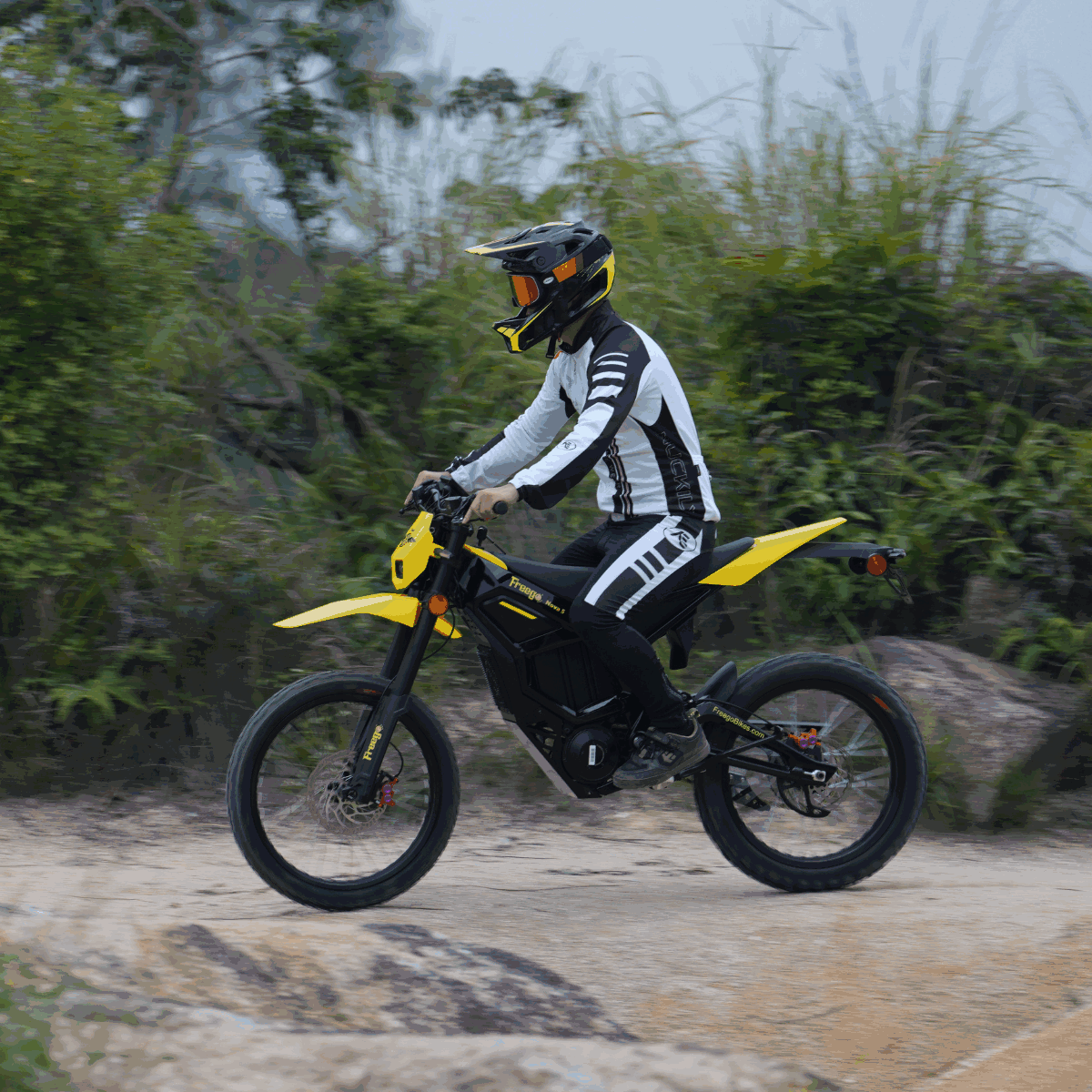 Rider on Freego Nova 5 electric dirt bike riding past dense greenery on an off-road trail.