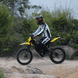 Rider on Freego Nova 5 electric dirt bike riding past dense greenery on an off-road trail.