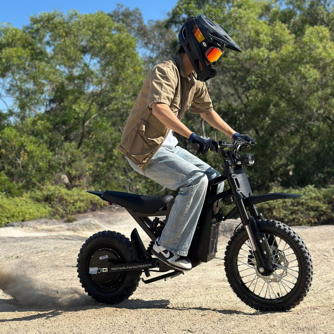 Helmeted rider on Freego X0 mini e-bike, kicking up dust while moving on gravel terrain, tree backdrop.