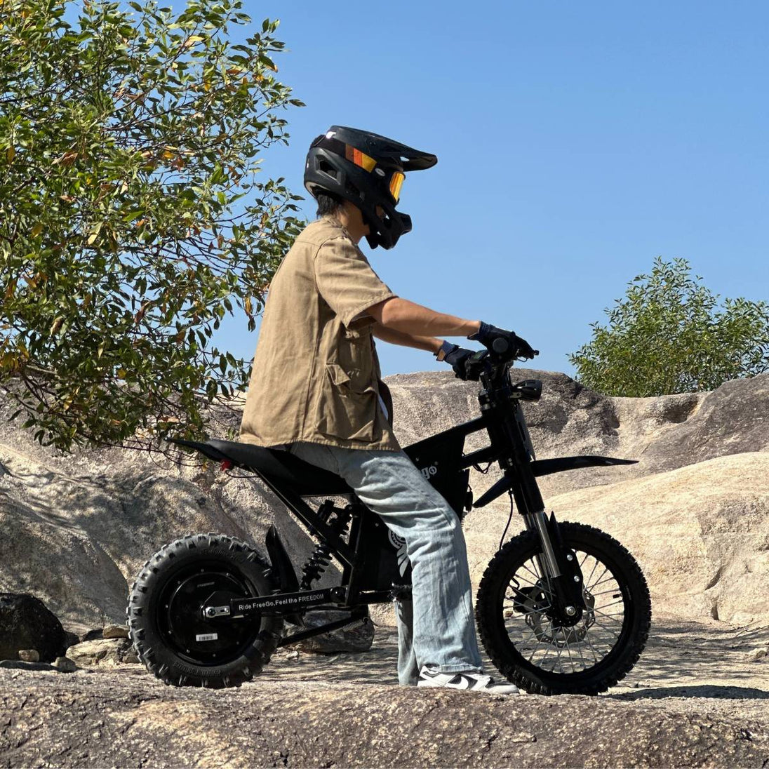 Helmeted rider seated on Freego X0 electric mini bike, positioned on sunlit rocky terrain with trees in the background.