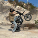 Helmeted rider doing a wheelie on a Freego-branded X0 electric mini dirt bike, set on sunlit rocky terrain.