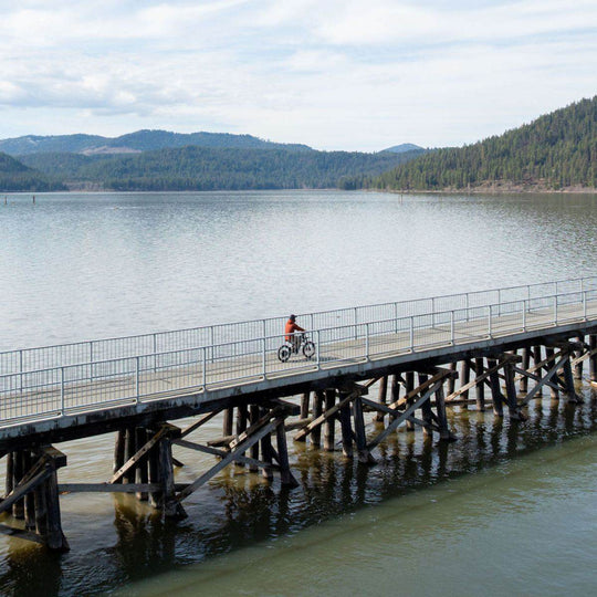Person riding a Freego X2 Pro dirt ebike on a wooden pier extending over a lake with mountains in the background.