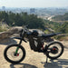 Freego X3 electric dirt bike (with pedals) parked on a rocky hill, city skyline and forested backdrop visible; brand logos and rugged tires on display.