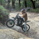 Freego X3 electric dirt bike navigates a rocky gully in a forested area; helmeted rider balances the bike, with ferns and trees lining the uneven trail.